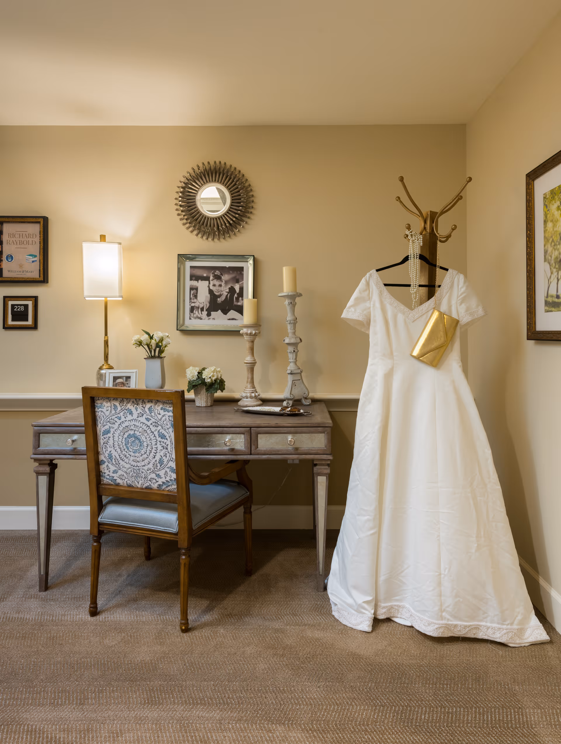 A furnished interior corner with a wooden desk and chair, decorative lamp and candles, framed photos on the wall, and a white dress hanging on a coat rack.
