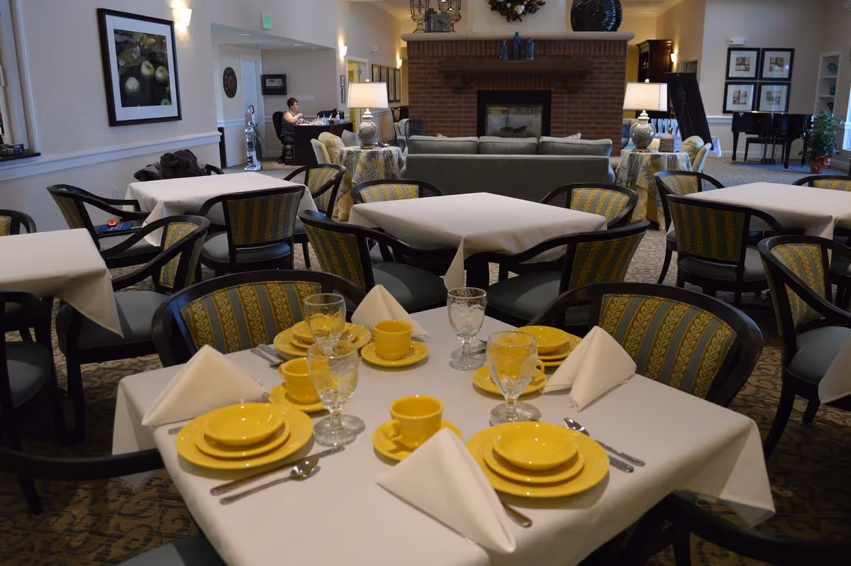 A dining room in a senior living facility with tables covered in white tablecloths, set with yellow plates, cups, and clear glasses. The chairs have patterned upholstery. In the background, there is a seating area with sofas and a brick fireplace, along with a piano and framed artwork on the walls.