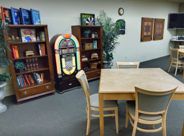 A senior living common room with a table and chairs, bookshelves, a retro jukebox, potted plants, and a wall-mounted TV.