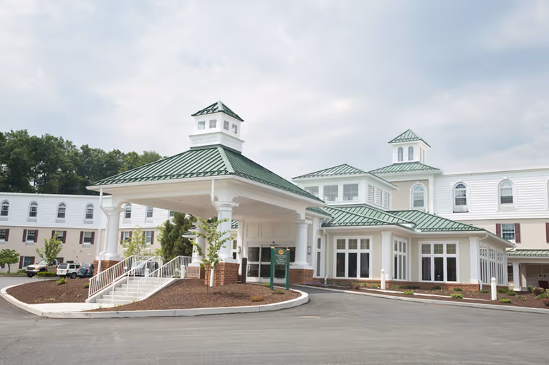 Exterior view of a large, white retirement community building with green metal roofs and cupolas. The building has a covered entrance with stairs and a ramp, surrounded by landscaped areas and a paved driveway.