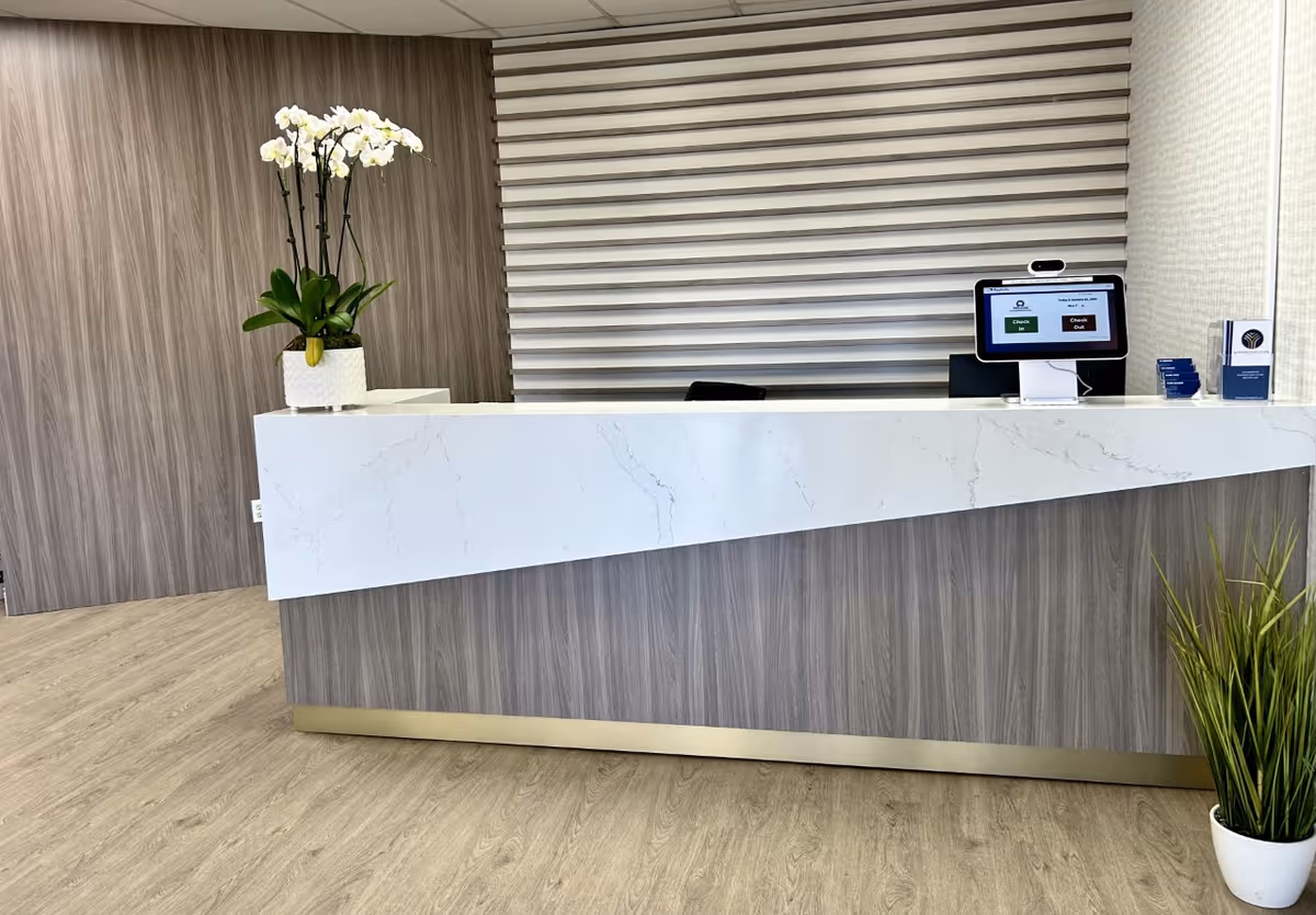 Reception desk area with a white marble-front counter, computer kiosk, orchid plant, and modern wood-paneled walls.