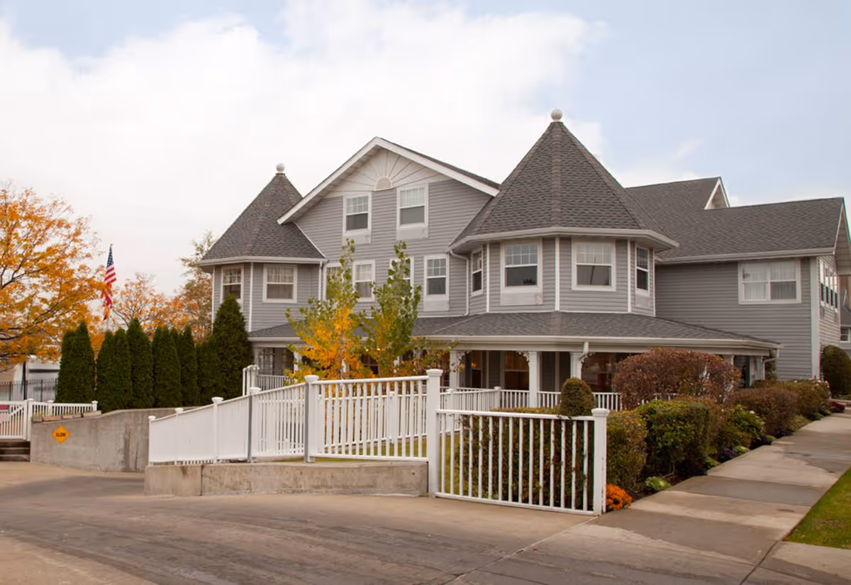 Exterior view of a large gray senior living facility building with multiple windows and two turret-like roof structures. The building is surrounded by a white fence, landscaped bushes, and trees with autumn foliage. An American flag is visible on the left side near the building.
