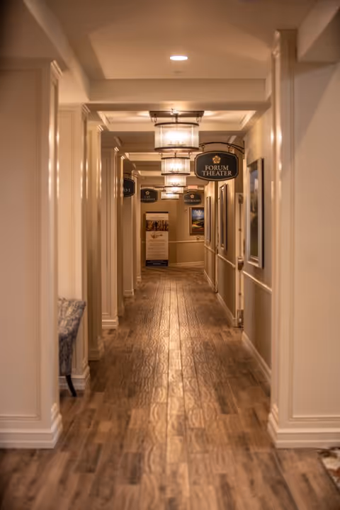 A well-lit hallway with wooden flooring and cream-colored walls, featuring multiple hanging signs including one for the Forum Theater. The hallway has framed pictures on the walls and ceiling lights evenly spaced along the corridor.