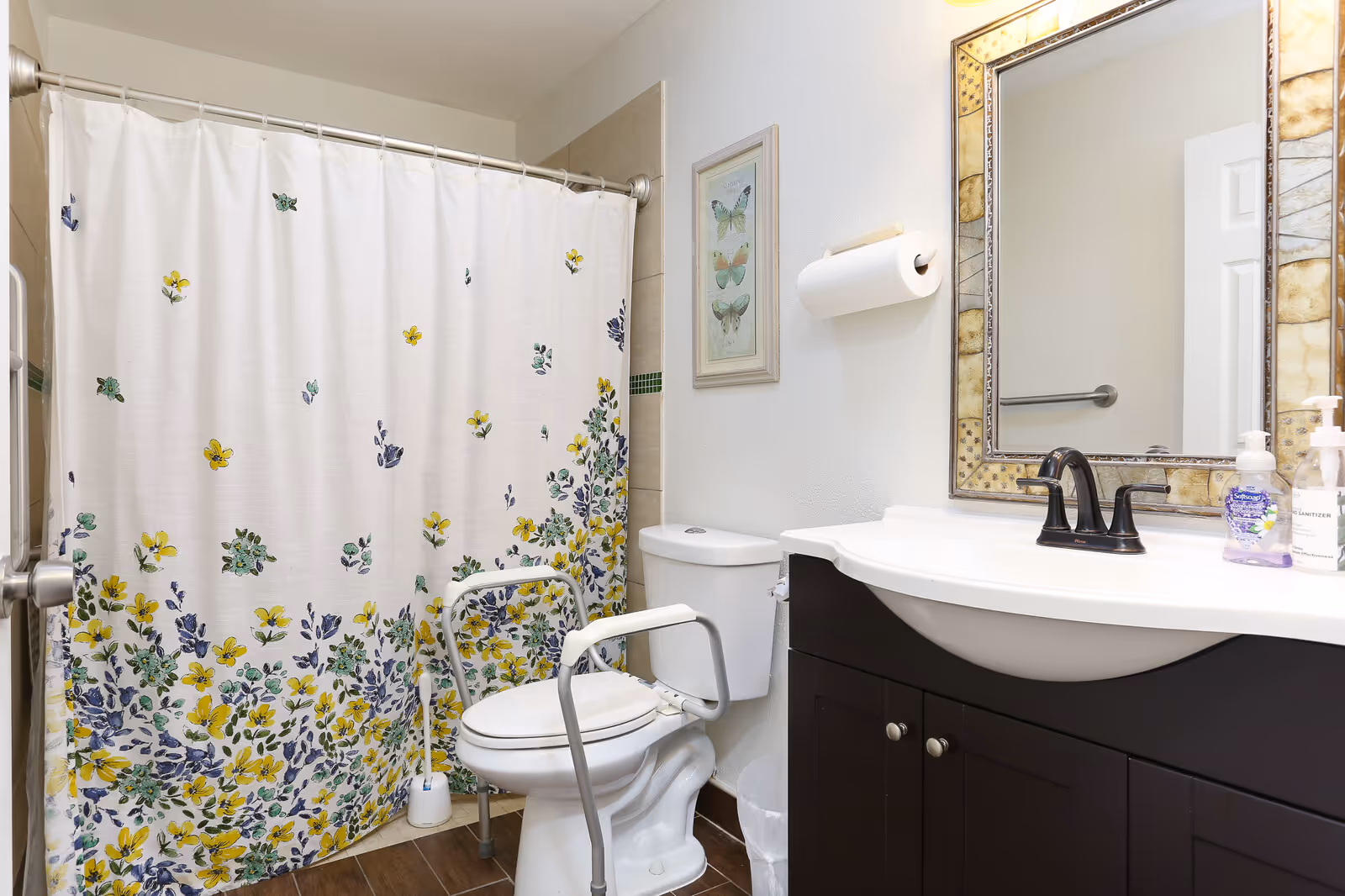 A bathroom with a white toilet equipped with safety rails, a white shower curtain decorated with yellow and purple flowers, a dark wood vanity with a white sink, a decorative framed mirror above the sink, a wall-mounted toilet paper holder, and hand soap and sanitizer bottles on the sink counter.