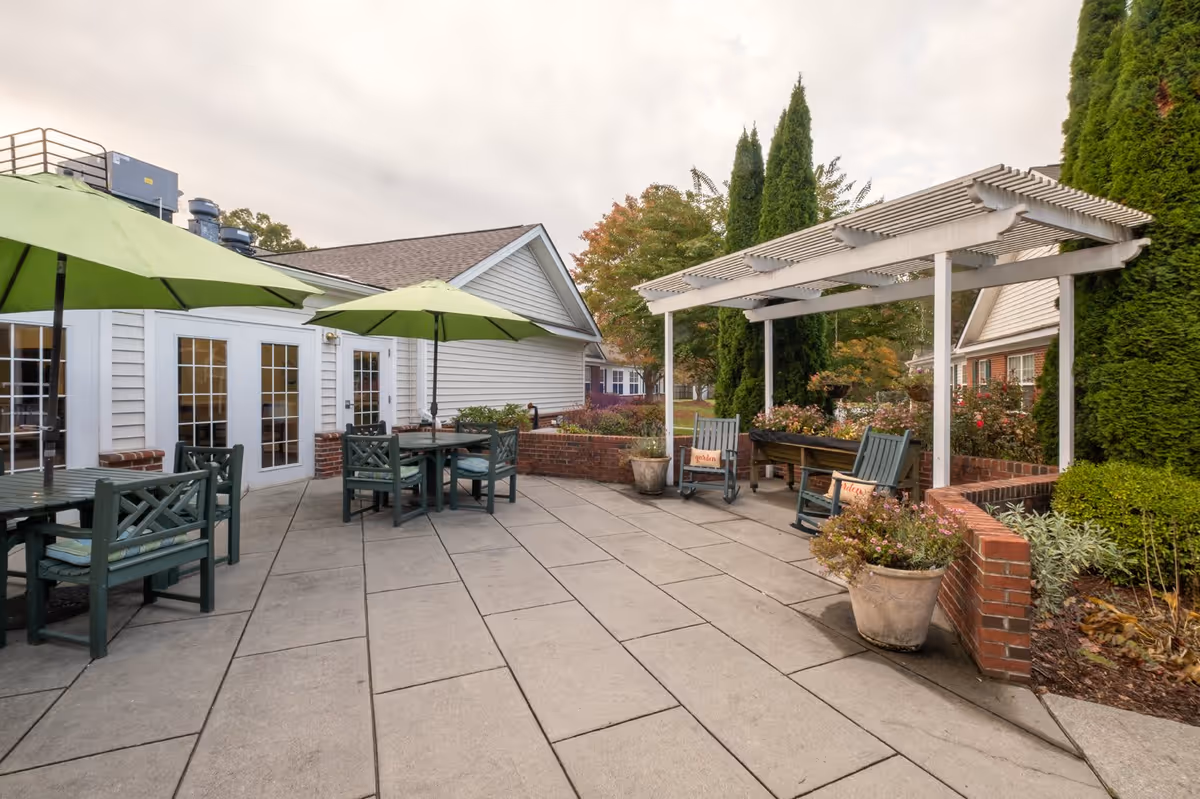 Outdoor patio area at Carolina Reserve of Laurel Park with green umbrellas shading tables and chairs, a white pergola with rocking chairs underneath, potted plants, and surrounding greenery including tall trees and bushes.