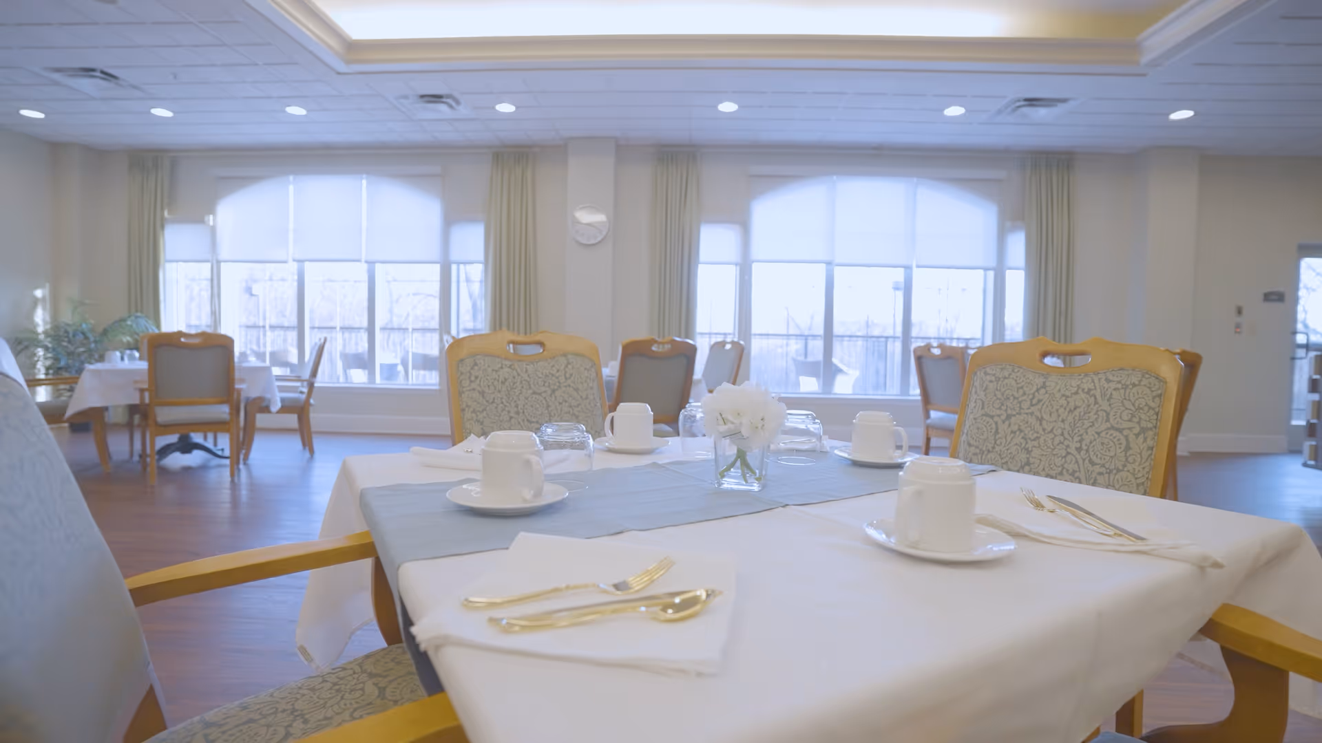 Bright dining room with set tables, upholstered chairs, white tablecloths, place settings, and a small floral centerpiece.