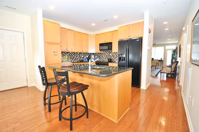 A modern kitchen with wooden cabinets, a black granite countertop island with a sink, and two black bar stools. The kitchen features a black refrigerator, a black microwave, and a tiled backsplash with a mosaic pattern. The floor is wooden, and the kitchen opens into a hallway leading to a living area with chairs and a table.