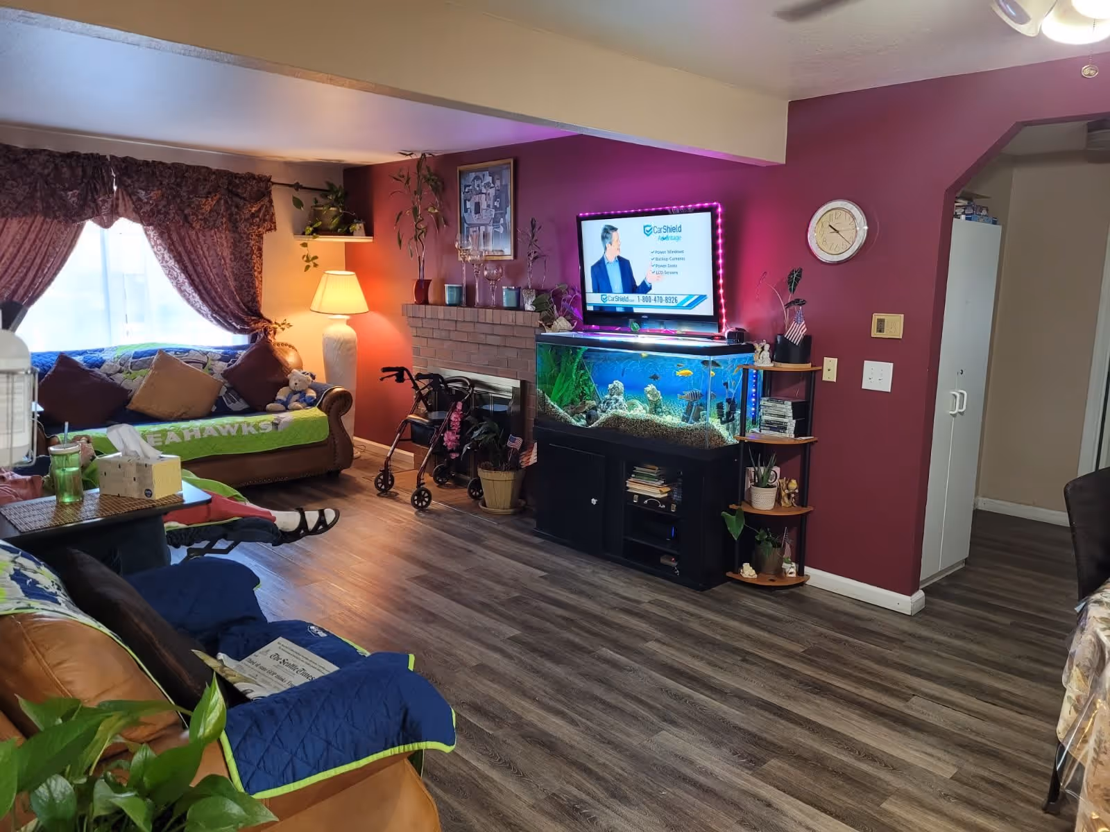 A cozy living room with two sofas covered in blankets and pillows, a large window with patterned curtains, a floor lamp, a walker, a brick fireplace, a large fish tank with colorful fish, a TV mounted above the fish tank with purple LED lights around it, a wall clock, and a small corner shelf with plants and decorative items.