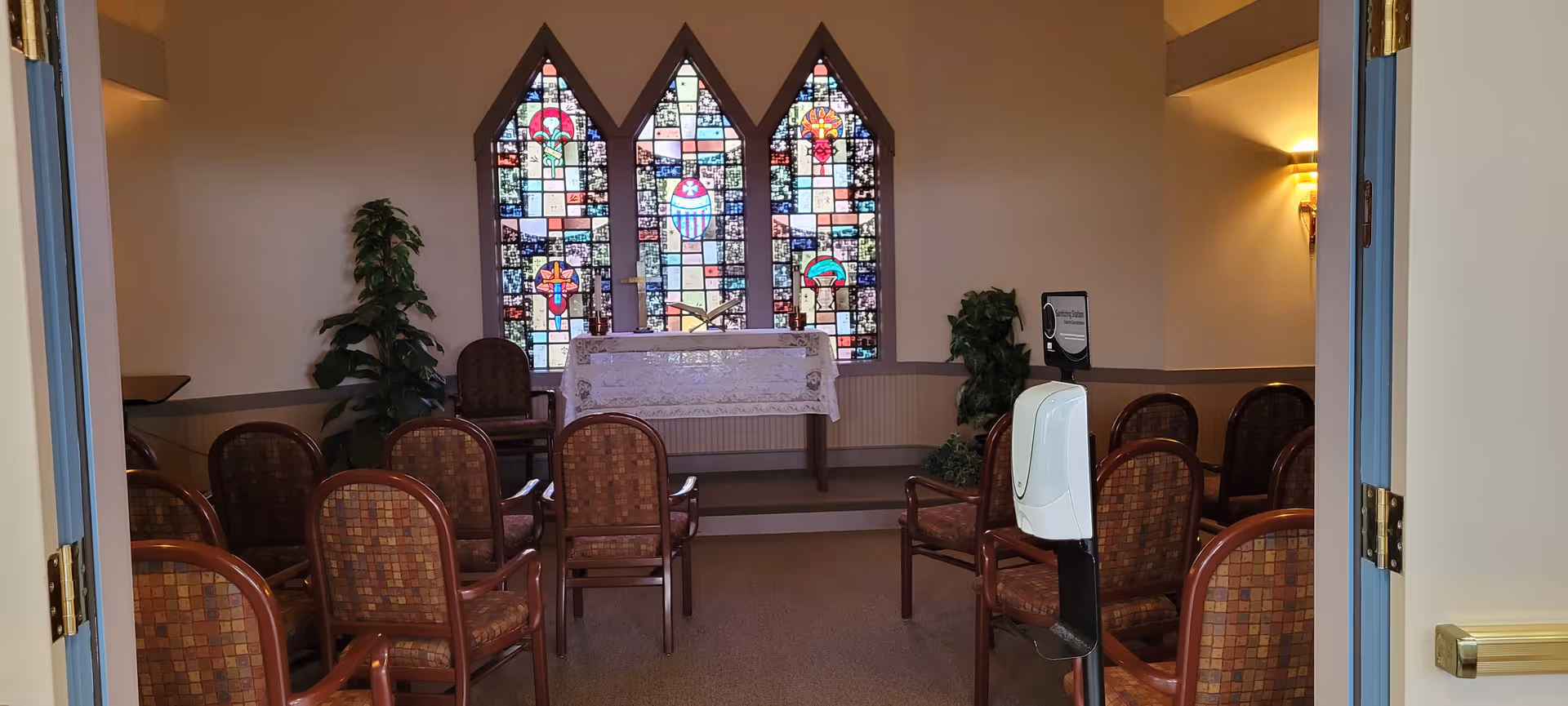 Small chapel room with rows of chairs facing an altar beneath three stained-glass windows.