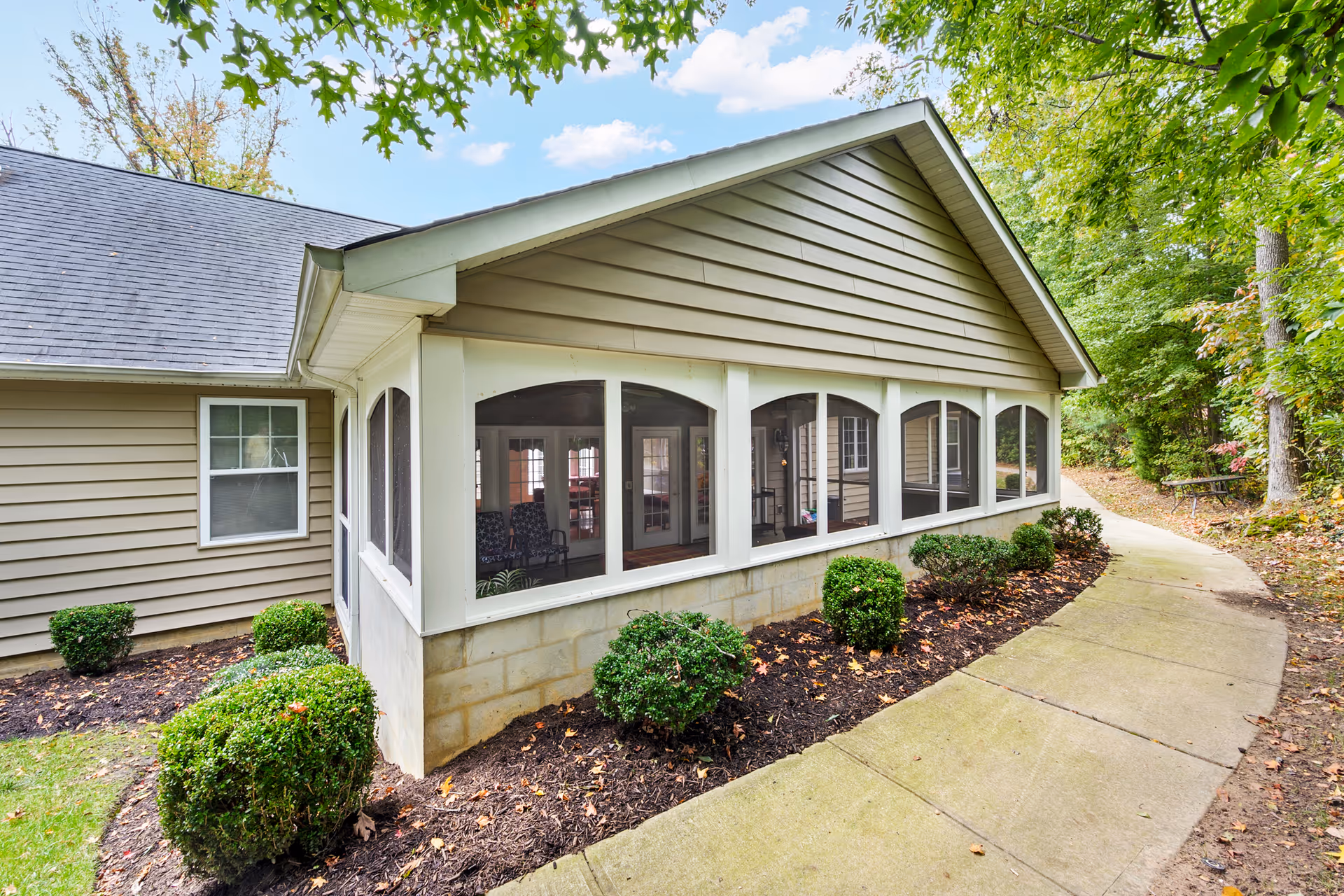 Exterior view of a single-story building with beige siding and a screened-in porch. The porch has multiple large windows and is surrounded by neatly trimmed bushes and a mulched garden bed. A curved concrete walkway runs alongside the building, with trees and greenery in the background under a partly cloudy sky.