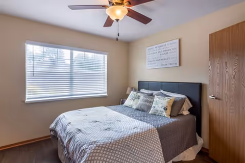 Bright bedroom with a bed dressed in patterned bedding, a ceiling fan, a large window with blinds, and wall art.