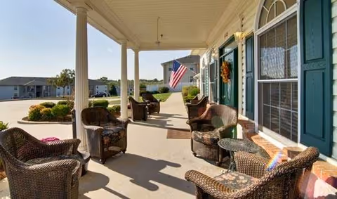 Covered front porch of a senior living facility with wicker chairs and tables, an American flag, and landscaped shrubs.
