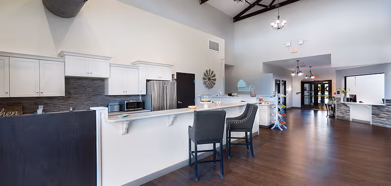 Spacious assisted-living common area with a long kitchen counter, two bar stools, and a reception area under high ceilings.