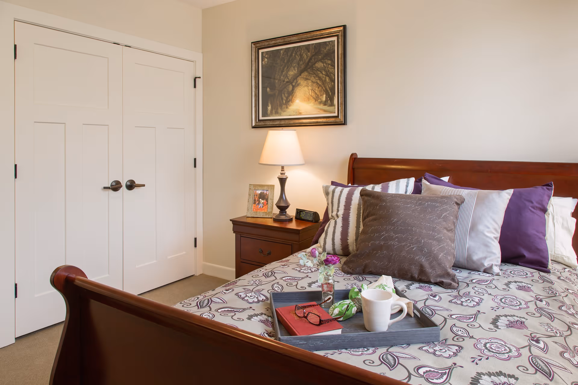 A cozy bedroom with a wooden bed frame and a bed covered in a floral patterned bedspread. The bed has several decorative pillows in shades of purple, gray, and white. On the bed is a tray holding a white coffee mug, a red book, a pair of glasses, and a small vase with flowers. Next to the bed is a wooden nightstand with a lamp, a framed photo, and a digital clock. Above the nightstand hangs a framed picture of a tree-lined path. There is a closed white double-door closet on the left side of the room.