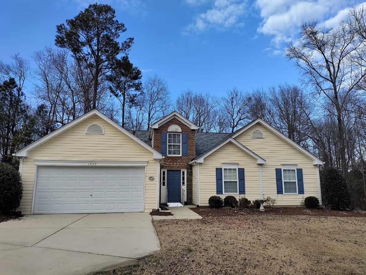 Front exterior view of a single-story house with beige siding, a two-car garage, blue shutters, and a blue front door. The house is surrounded by leafless trees and a dry lawn under a partly cloudy blue sky.