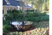 Outdoor garden area at Forest Haven Nursing & Rehabilitation Center, LLC featuring a large reflective water bowl surrounded by green plants and flowers, with part of the building visible in the background.