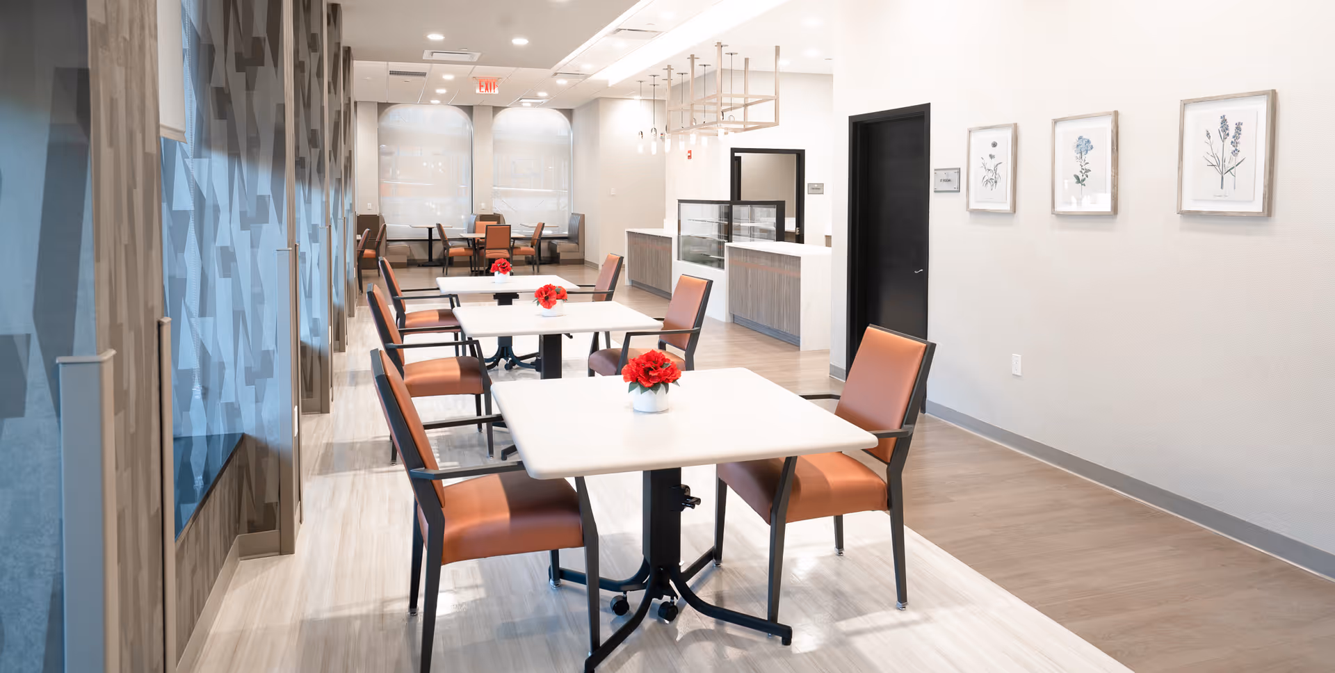 A bright and clean dining area in a senior living facility with several square tables, each adorned with a small red flower centerpiece. The chairs have brown cushions and black frames. The room features light-colored walls with three framed botanical prints, a patterned glass wall on the left, and a counter area in the background with modern lighting fixtures.