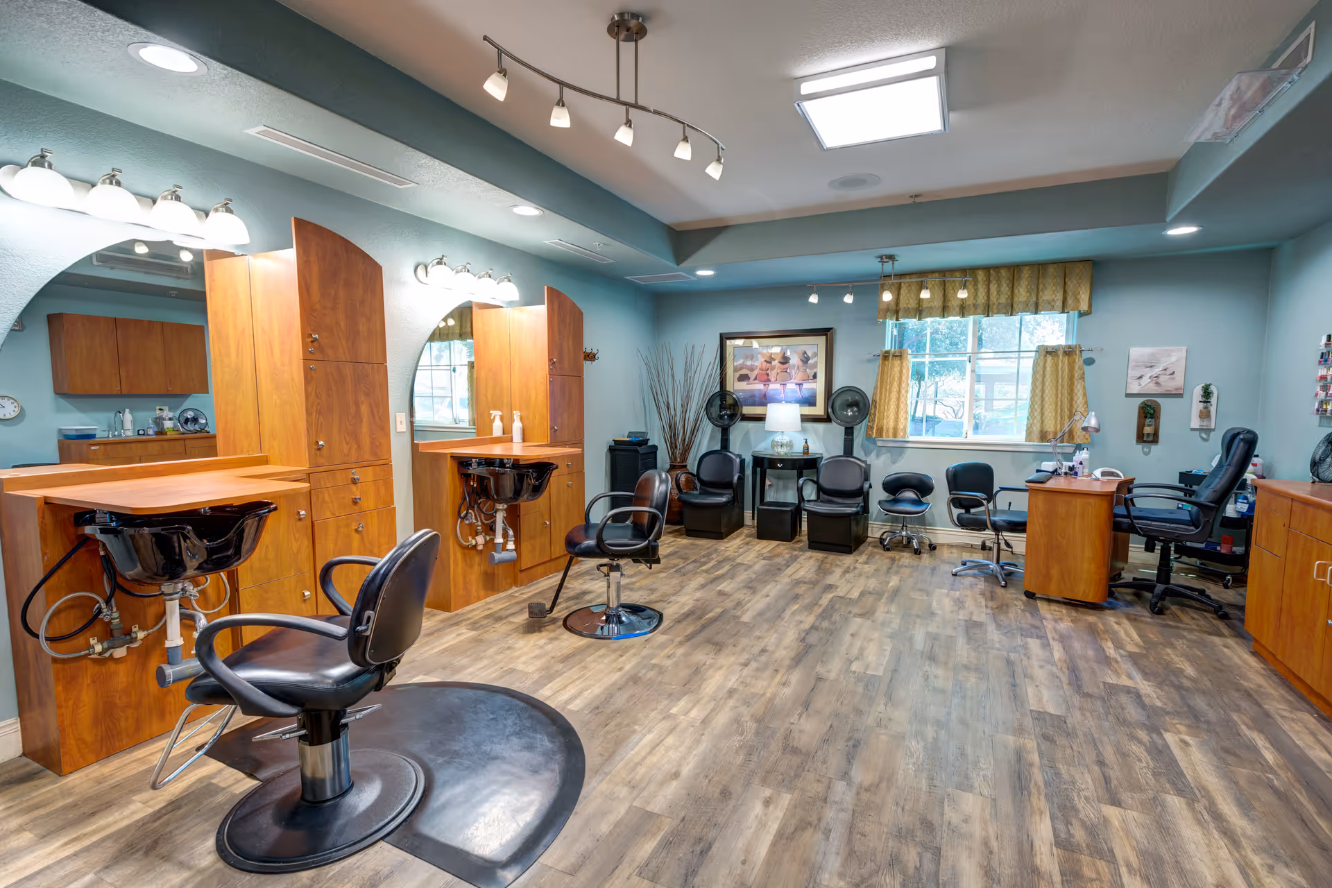 Interior view of a senior living facility's hair salon with wooden cabinetry, two black salon chairs in front of wash basins, multiple black chairs along the wall, a desk with an office chair, and a window with yellow curtains letting in natural light.