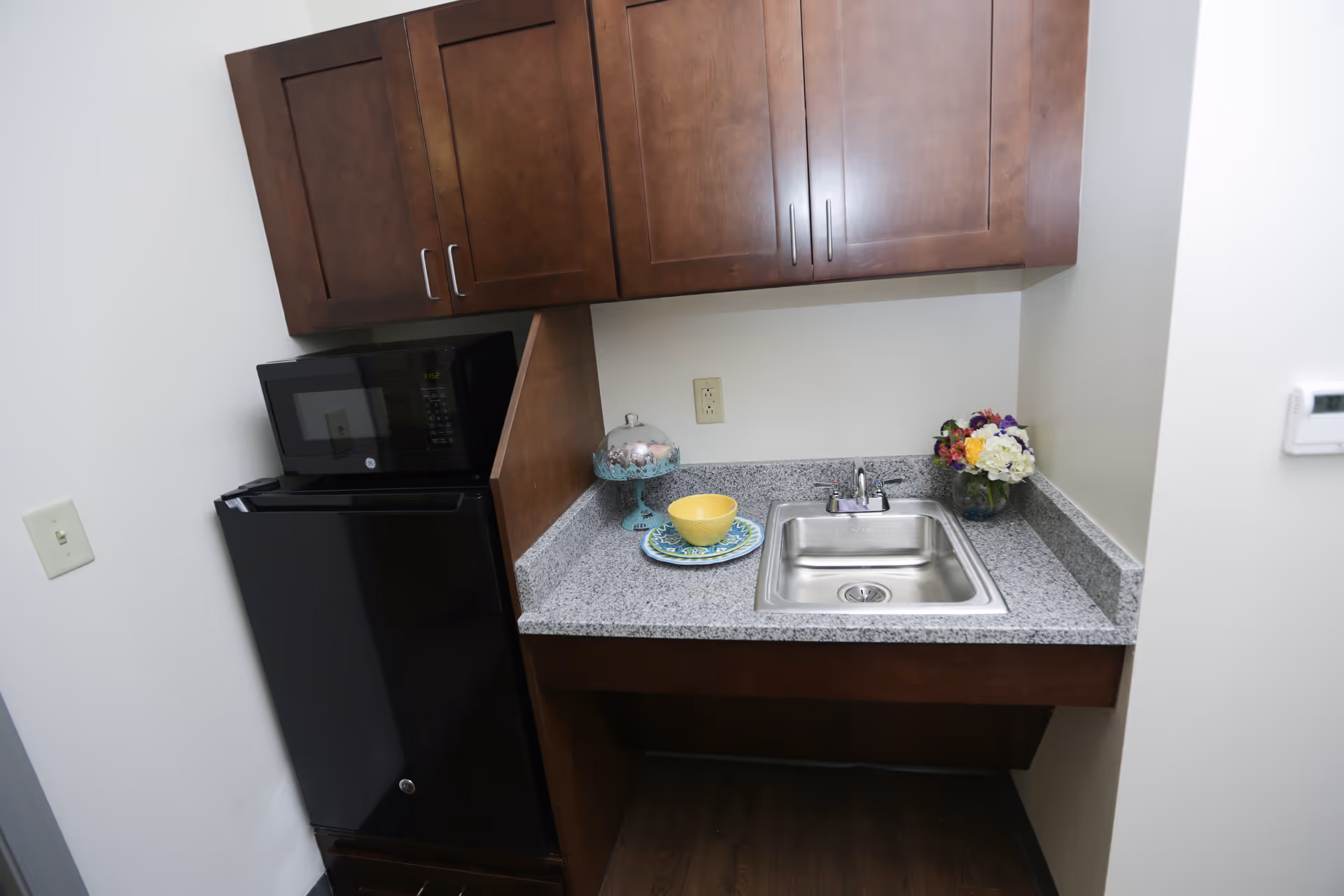 Small kitchenette area with a black mini refrigerator and microwave stacked on top, wooden cabinets above, a granite countertop with a stainless steel sink, a yellow bowl on a decorative plate, a glass cake stand with a cover, and a vase of colorful flowers on the right side.