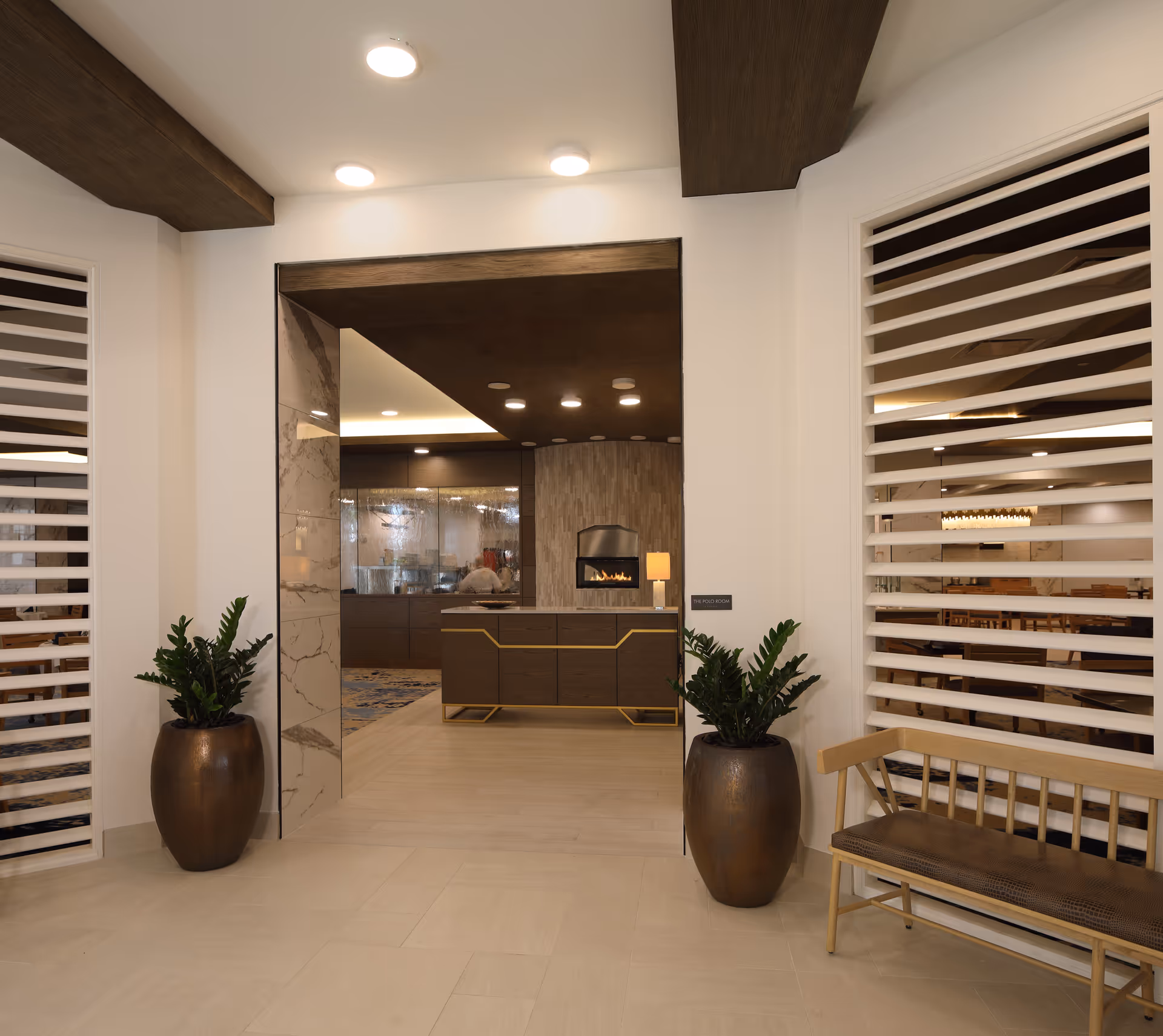A modern lobby/reception area with a front desk, fireplace, potted plants, and a wooden bench.