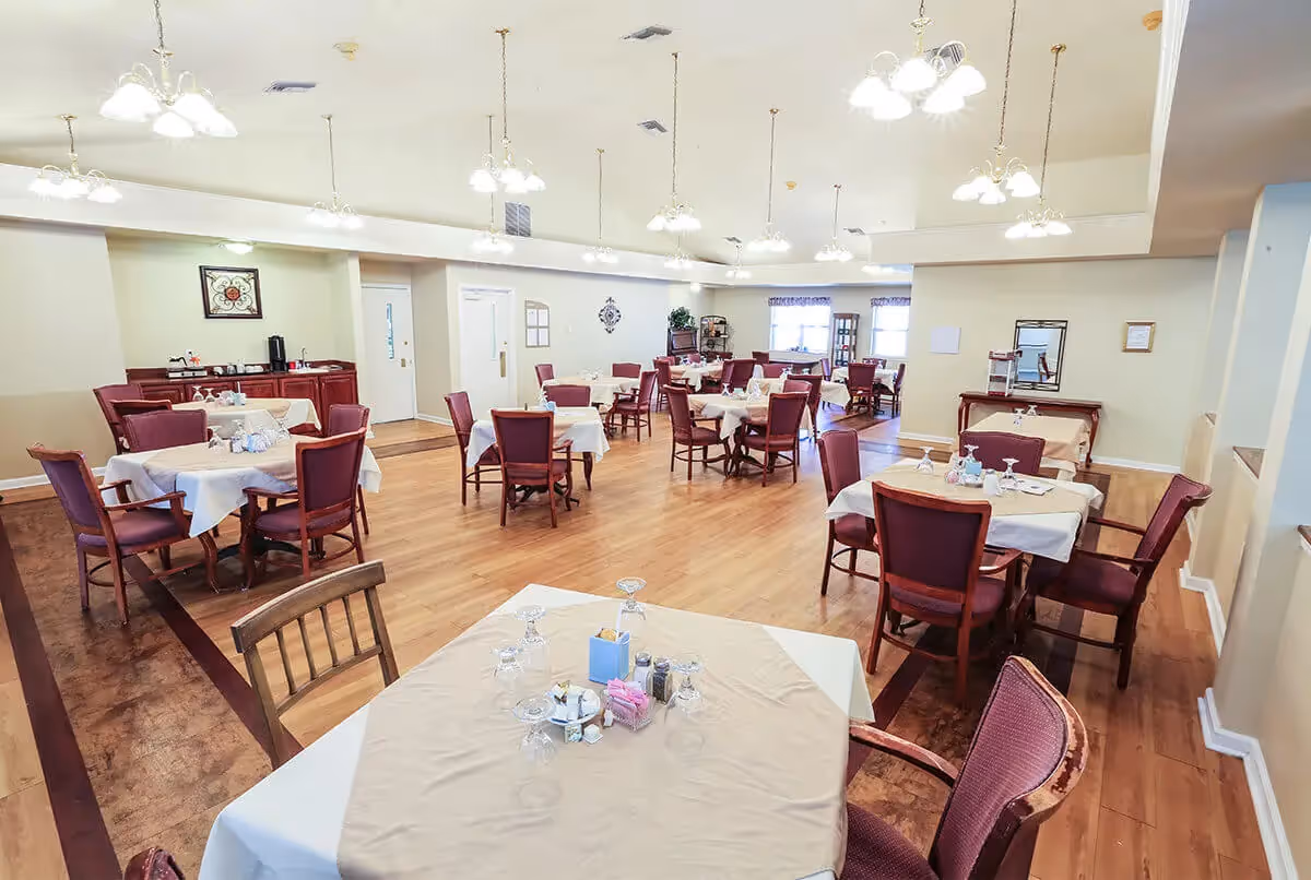 A spacious dining room with multiple tables covered in beige tablecloths and set with glasses and condiments. The room has wooden floors, cream-colored walls, and several hanging light fixtures. Chairs with red upholstery surround the tables, and there are windows letting in natural light in the background.