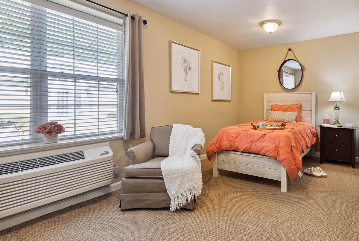 A cozy bedroom with a single bed dressed in an orange comforter and a decorative pillow that says 'Family'. Next to the bed is a dark wooden nightstand with a lamp, tissues, and a small clock. A round mirror hangs on the wall above the bed's headboard. There is a comfortable armchair with a white throw blanket draped over it near a window with blinds and beige curtains. A small vase with pink flowers sits on the windowsill. The walls are painted a warm beige color, and two framed botanical prints hang on the wall above the armchair.