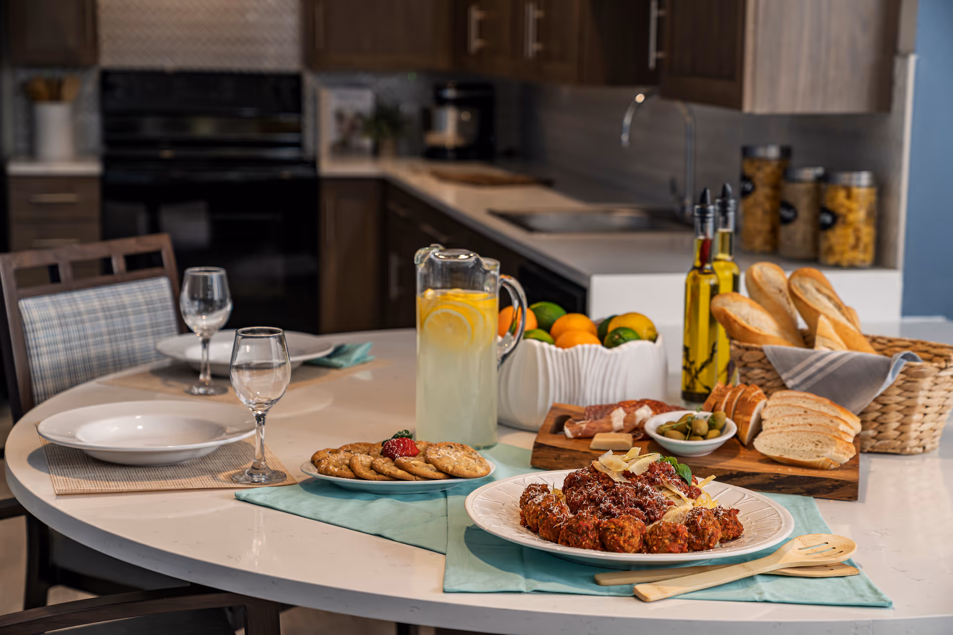A kitchen dining area with a round table set for two, featuring plates, wine glasses, and a pitcher of lemonade. On the table are plates of cookies, a dish of meatballs with sauce, a basket of bread, a bowl of olives, and bottles of olive oil. The background shows kitchen cabinets, a sink, and a stove.