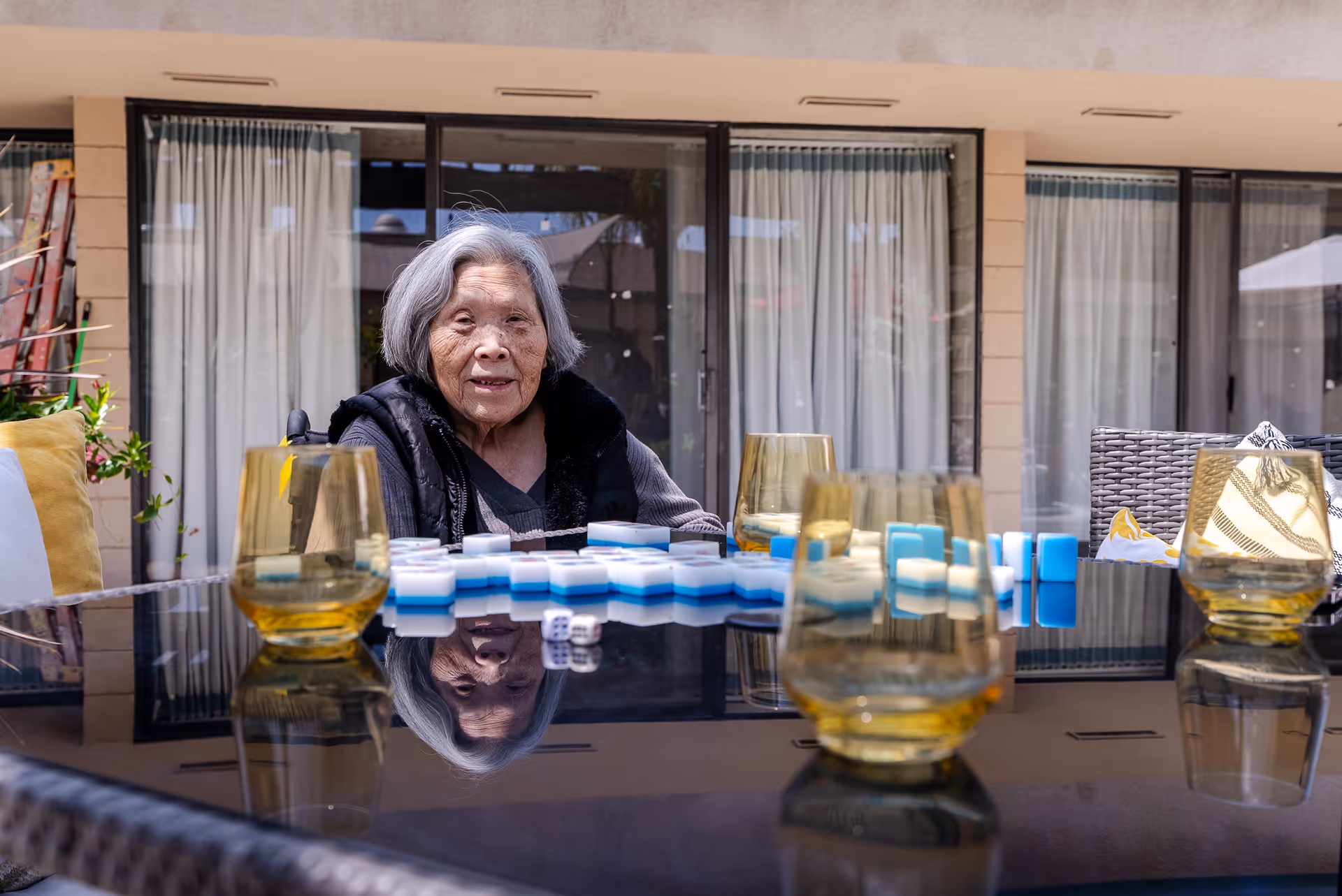 An elderly woman with gray hair sitting at a glass outdoor table with yellow-tinted glasses and blue and white game pieces, in front of a building with large windows and white curtains.