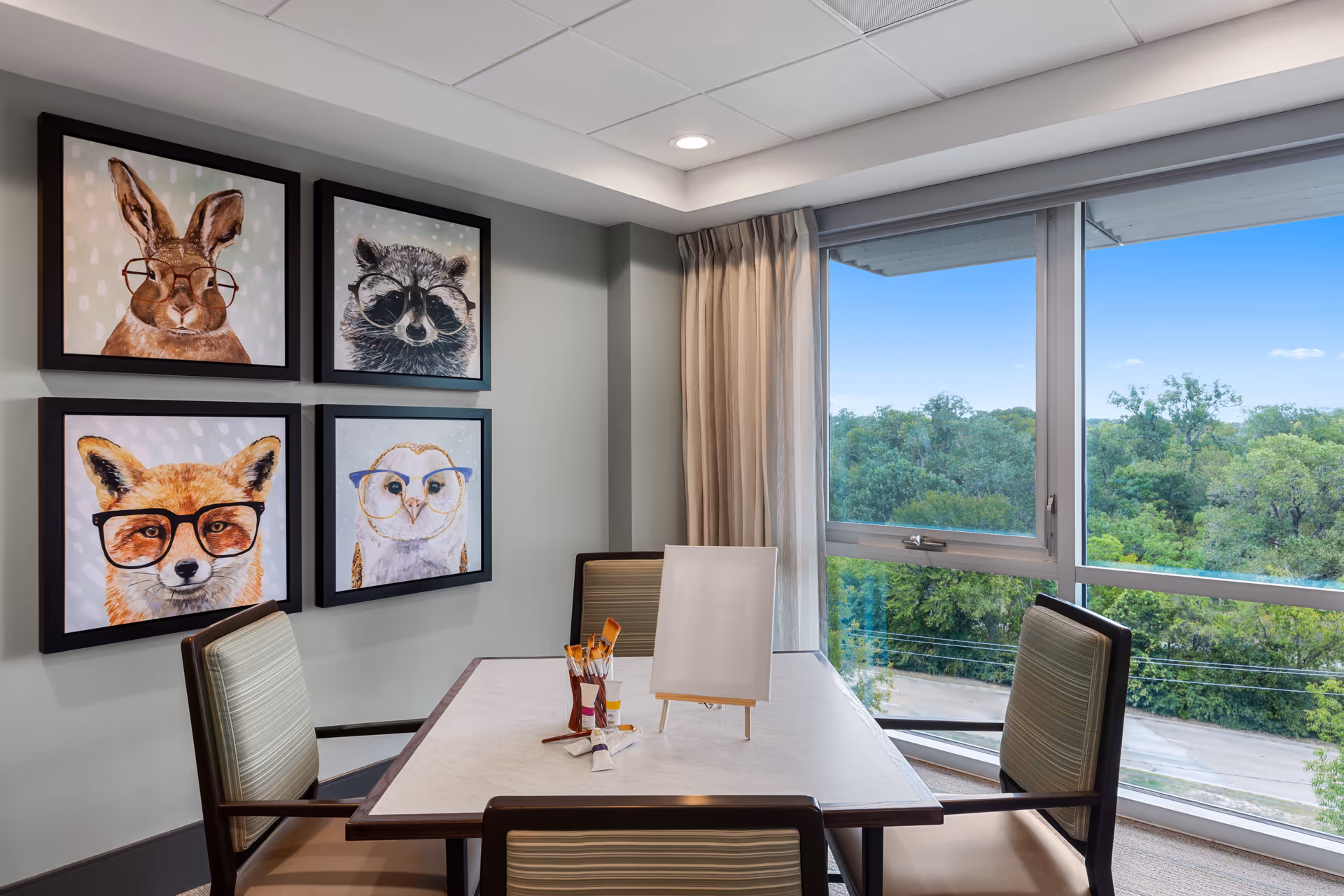 A small meeting or activity room with a square table surrounded by four chairs. On the table are paintbrushes, paint tubes, and a blank canvas on a small easel. The room has large windows with a view of green trees outside. On the wall are four framed animal portraits featuring a rabbit, raccoon, fox, and owl, each wearing glasses.