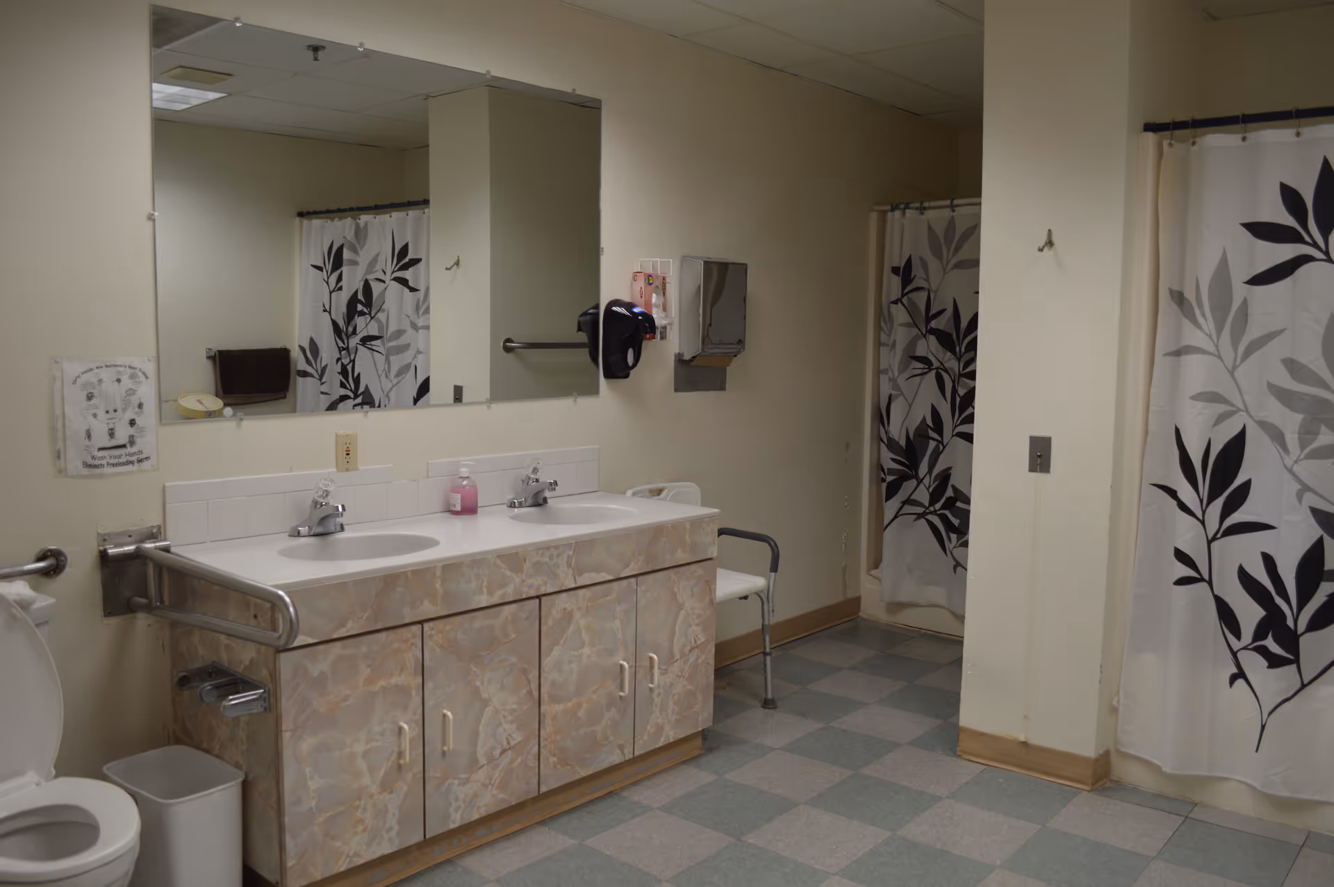 A bathroom with two sinks and a large mirror above them. There is a toilet with an open lid on the left side, a trash bin, and a grab bar next to the toilet. The shower area has a curtain with a black and gray leaf pattern. A white chair with armrests is placed near the shower. The walls are light-colored, and the floor has a checkered pattern of green and gray tiles.