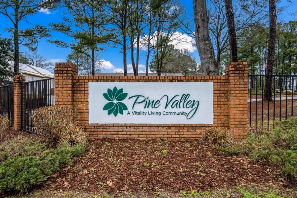 Brick sign at the entrance of Pine Valley Retirement Community with a green floral logo and the text 'Pine Valley A Vitality Living Community', surrounded by trees and landscaping under a blue sky with clouds.