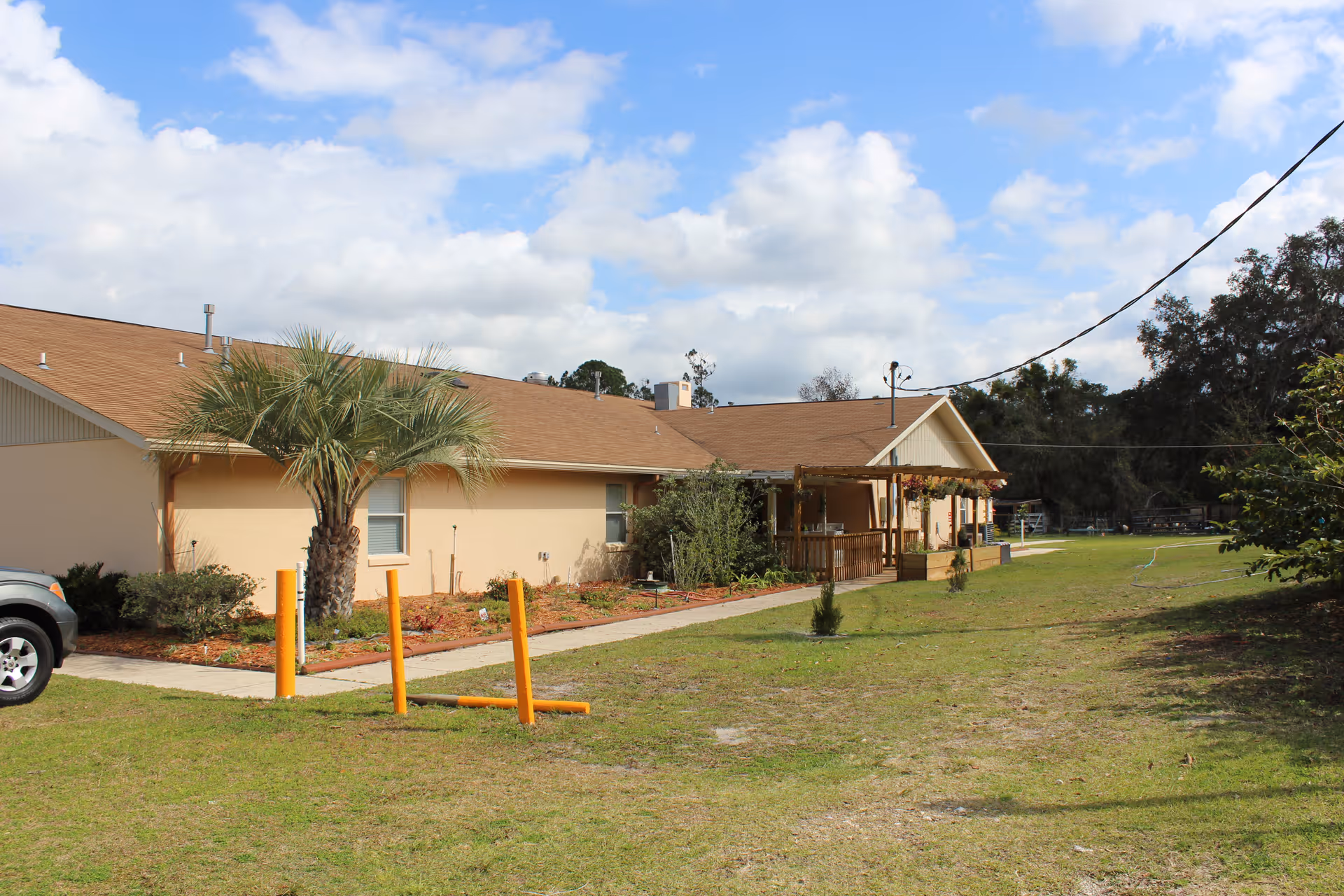 Single-story beige building with a front porch, palm tree, and grassy yard under a partly cloudy sky.
