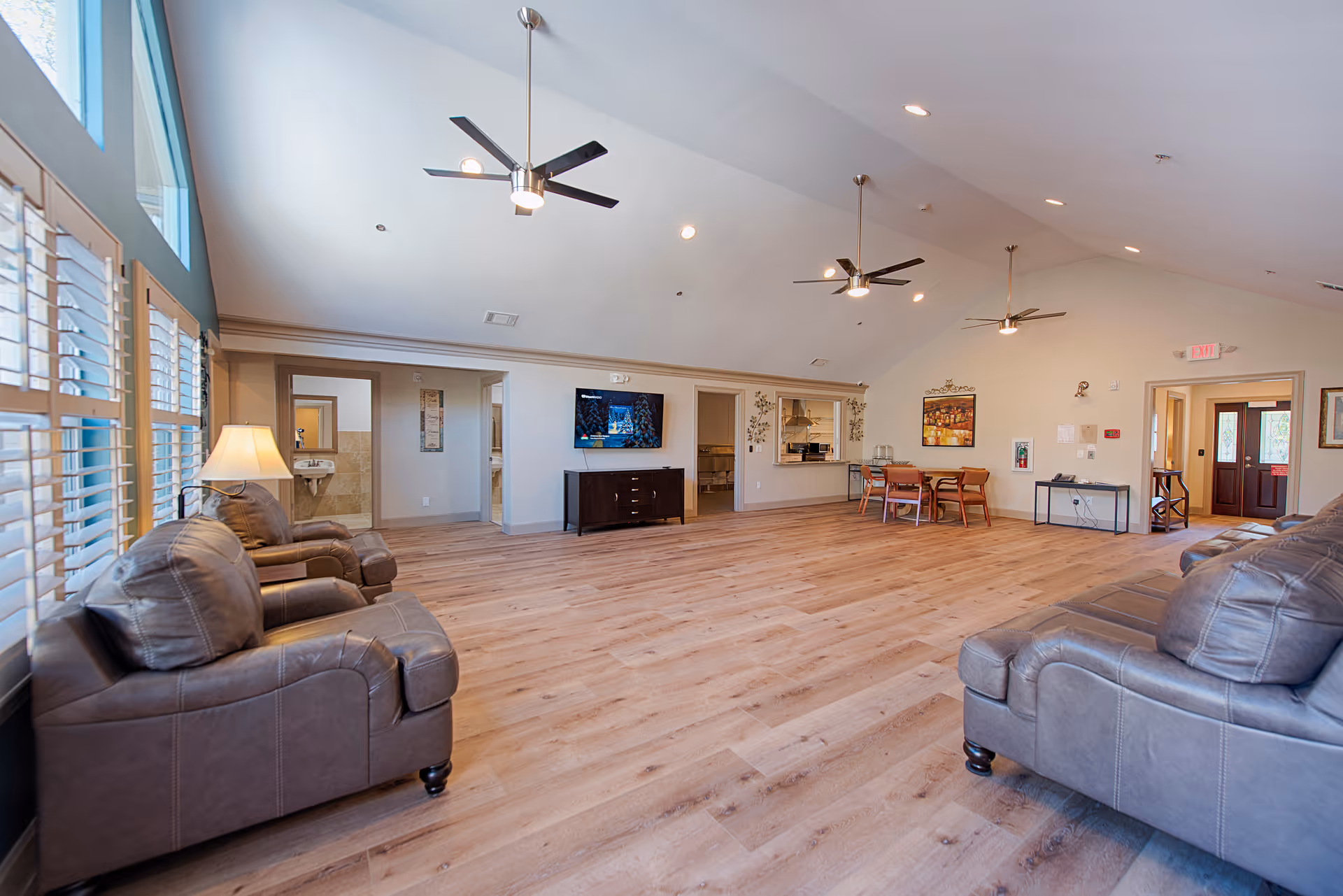 A spacious living room with high vaulted ceilings featuring three ceiling fans. The room has large windows with white shutters on the left side, allowing natural light to fill the space. There are two brown leather sofas facing each other on opposite sides of the room. A flat-screen TV is mounted on the far wall above a dark wooden cabinet. To the right, there is a small dining area with a table and four chairs. The floor is covered with light wood laminate, and the walls are painted in a neutral tone. The room has multiple doorways leading to other areas of the facility.