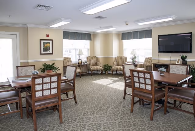 A bright and spacious common area in a senior living facility featuring two wooden tables with chairs, several armchairs arranged near windows with blue valances, a flat-screen TV mounted on the wall, and decorative plants and framed artwork.