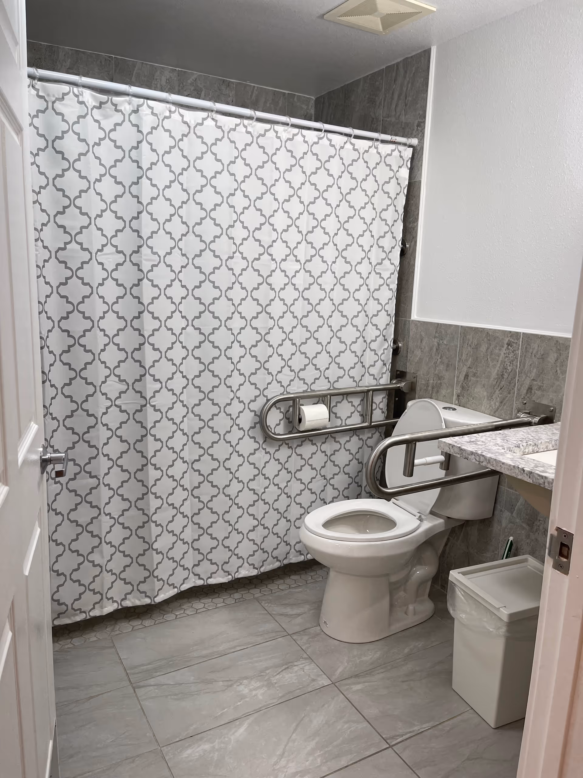A clean bathroom with a white toilet equipped with metal grab bars on both sides for accessibility. There is a white and gray patterned shower curtain covering the bathtub. The floor and lower walls are tiled in gray, and there is a white trash bin next to the toilet. The bathroom has a white door partially open.