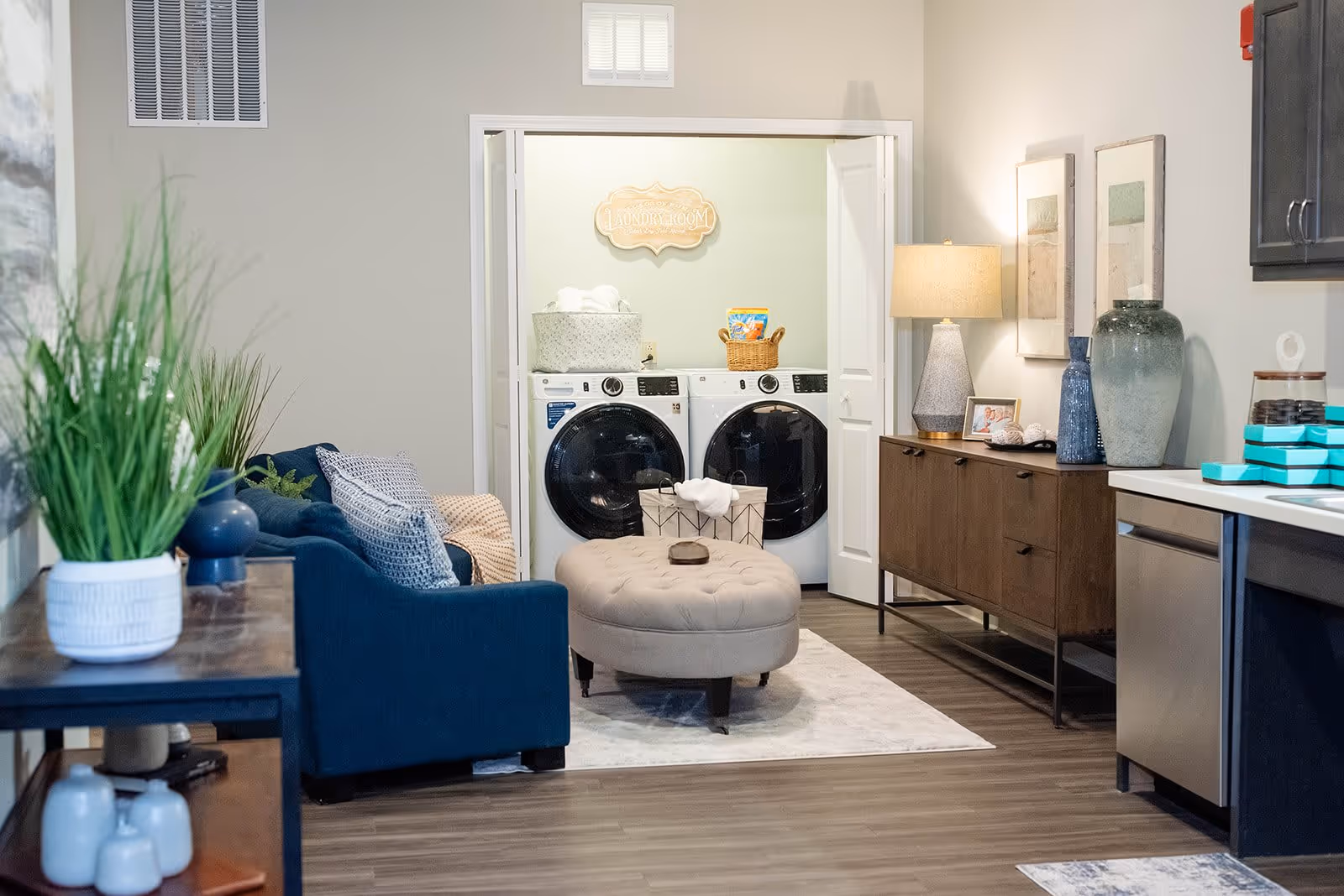 A cozy interior space featuring a blue sofa with cushions, a beige tufted ottoman on a light rug, and a wooden sideboard with decorative items and a lamp. In the background, there is an open laundry room with a front-loading washer and dryer, laundry baskets, and a decorative sign above the machines.