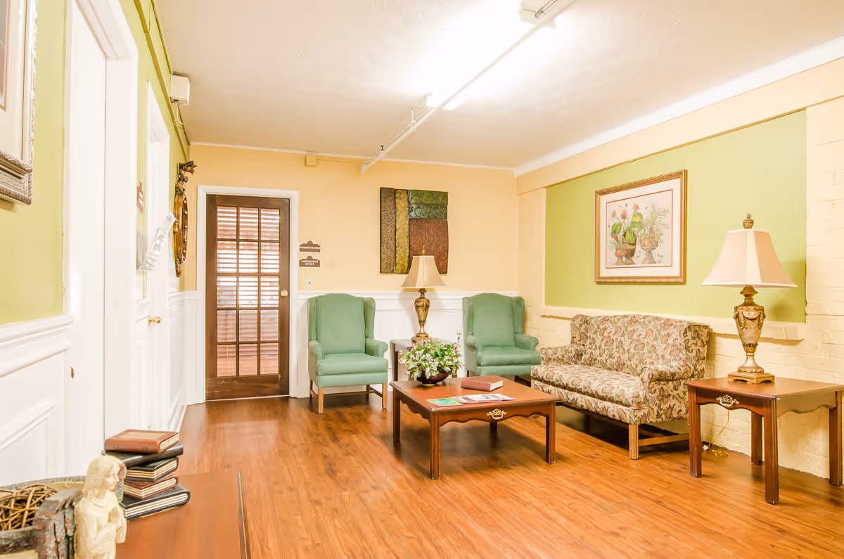 A cozy sitting area with wooden flooring, featuring two green armchairs, a floral-patterned sofa, a wooden coffee table with a plant and books, two side tables with lamps, and framed artwork on the walls. A wooden door with glass panels is visible in the background.