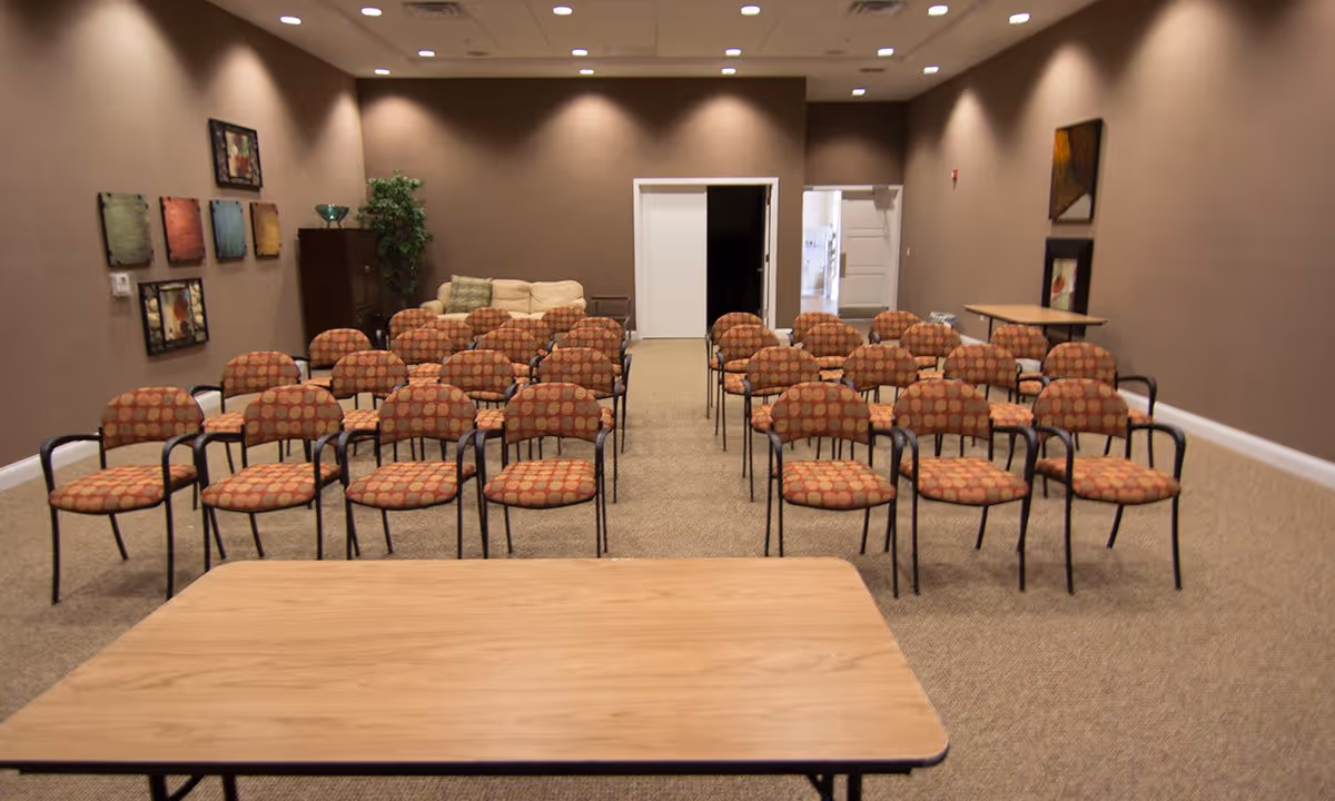 A conference or meeting room with rows of patterned chairs arranged facing a wooden table. The room has brown walls decorated with framed artwork, a beige carpet, a beige couch in the back corner, and two white doors at the far end.