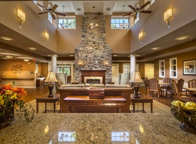 Spacious and well-lit senior living facility common area with a large stone fireplace in the center, surrounded by comfortable seating including sofas and armchairs. The room features high ceilings with ceiling fans and wall sconces, large windows letting in natural light, and wooden tables and chairs arranged for socializing or dining. Decorative plants and lamps add warmth to the inviting interior space.