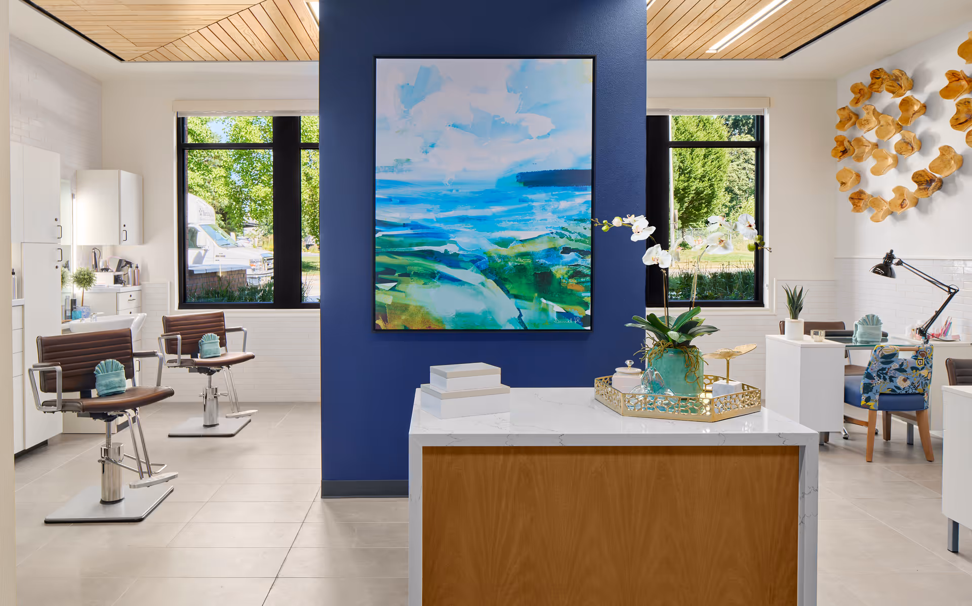 A bright and modern interior space featuring two brown salon chairs with teal towels on them, white cabinetry, and large windows showing greenery outside. A blue accent wall with a colorful abstract painting divides the room. In the foreground, a white countertop with a wooden base holds a decorative tray with a plant and other items. On the right side, there is a desk with a floral-patterned chair, a desk lamp, and wall art made of wooden pieces.