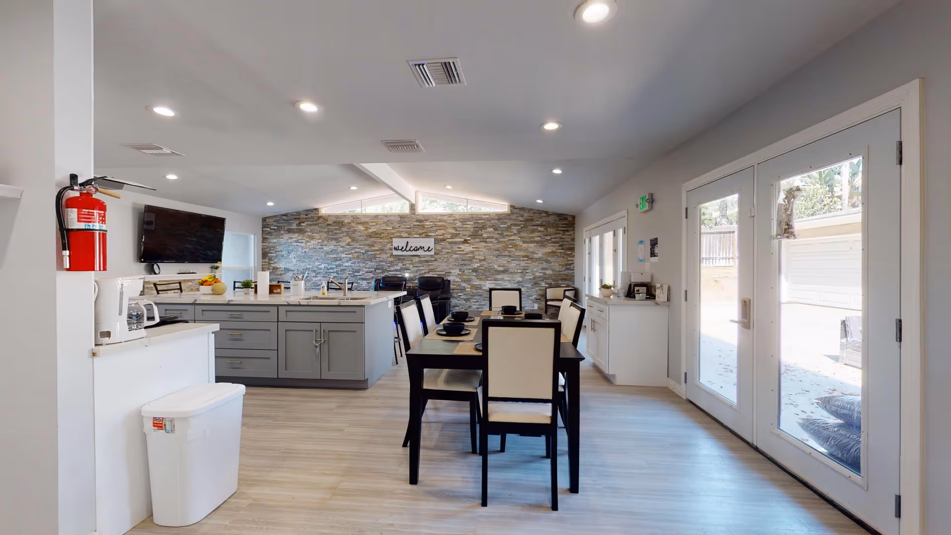 Bright communal dining and kitchen area with a central dining table, island, stone accent wall and double glass doors.