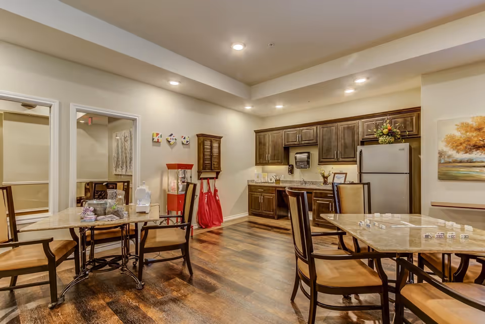 Interior view of a common area in Clear Fork Assisted Living and Memory Care featuring a kitchenette with dark wood cabinets, a refrigerator, and a countertop. There are two tables with chairs around them, one table has a game set up with dominoes. The room has wood flooring, recessed ceiling lights, and a painting of a tree on the wall.