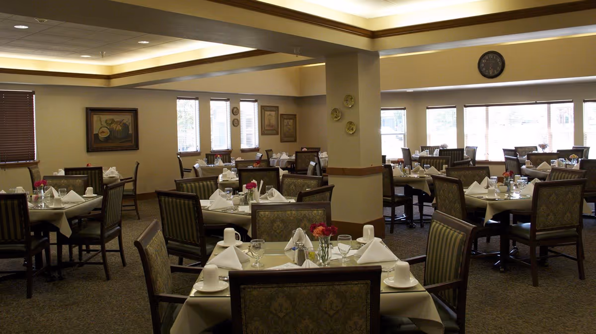 A dining room in a senior living facility with multiple tables set for meals. Each table has white tablecloths, folded napkins, glasses, cups, and small flower arrangements. The room has beige walls, several windows with blinds, framed artwork, and a clock on the wall.