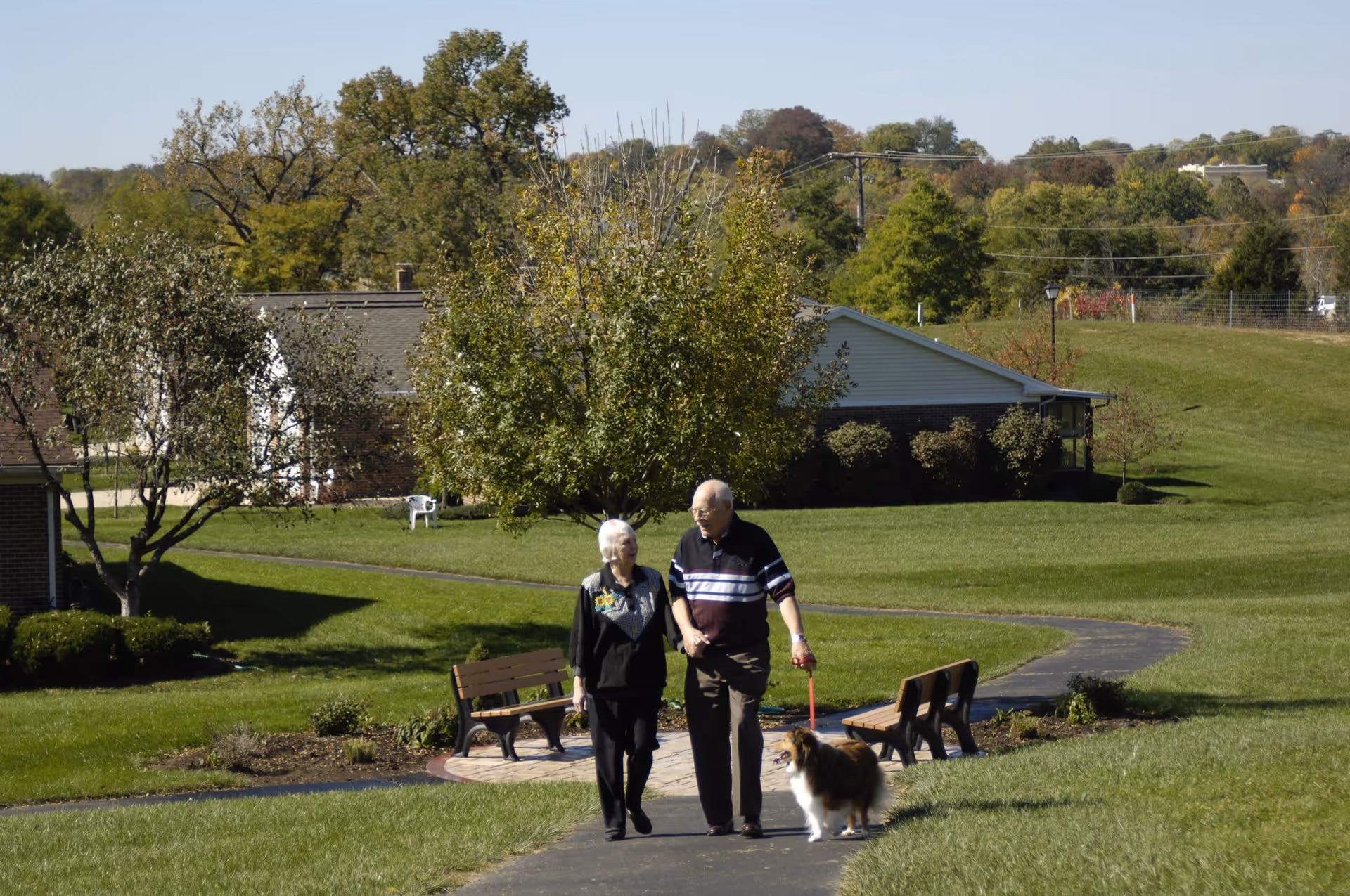 An elderly couple walking hand in hand on a paved path in a green outdoor area with benches and trees, accompanied by a small dog on a leash. Residential buildings and trees are visible in the background under a clear sky.