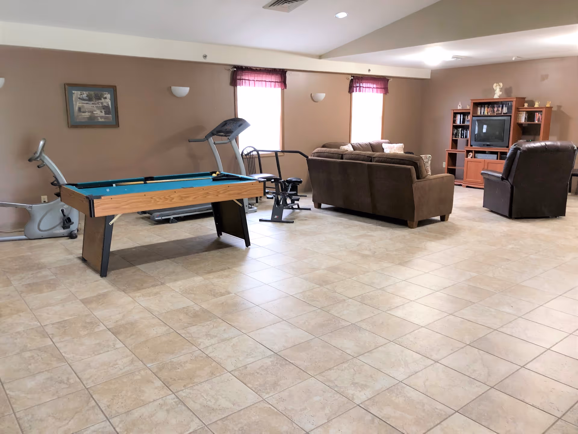 A spacious room with beige tiled flooring and light brown walls featuring a pool table, exercise equipment including a stationary bike and treadmill, and a seating area with a brown couch, a recliner, and a wooden entertainment center with a TV and shelves.