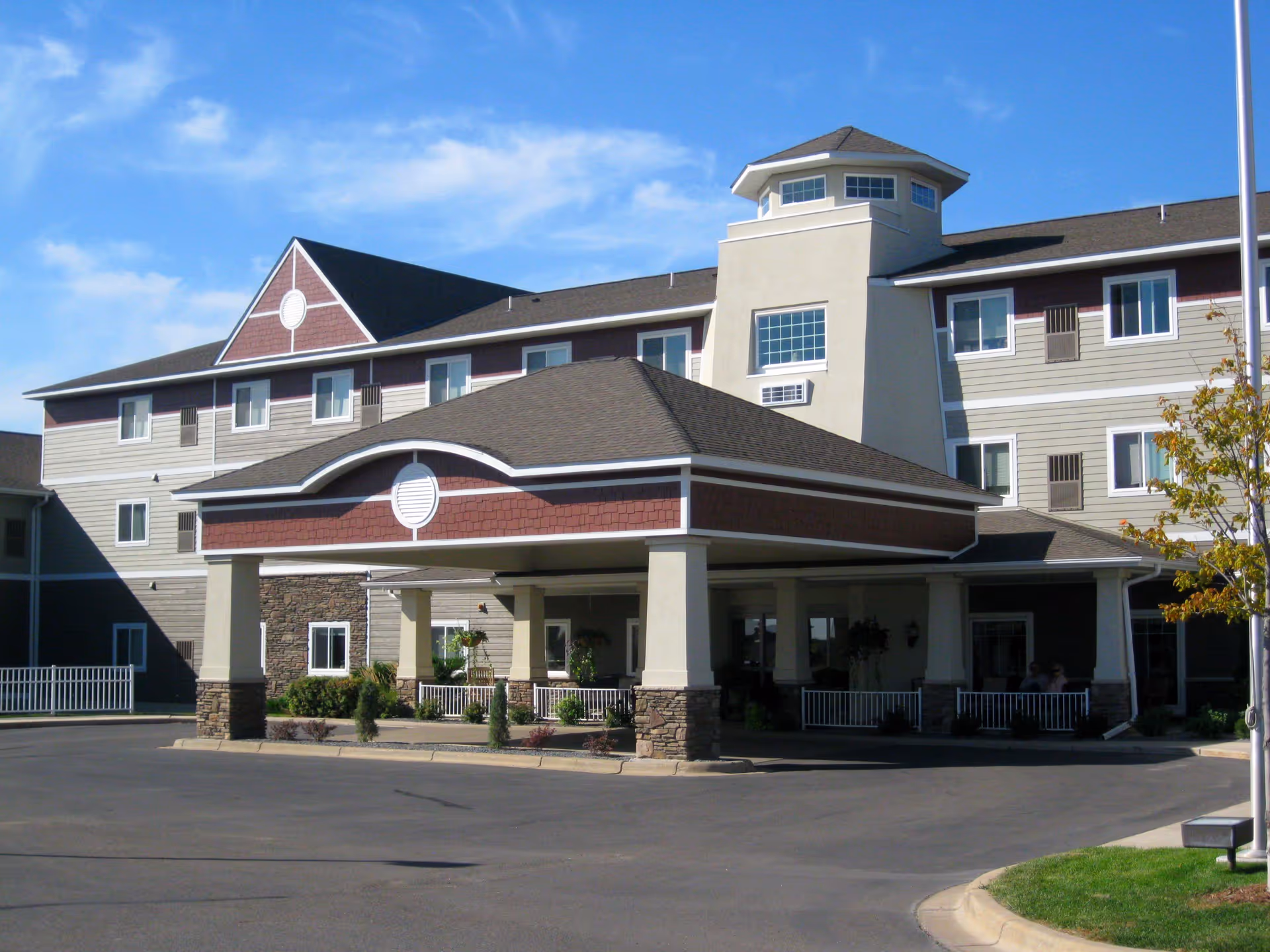 Exterior view of a multi-story senior living facility building with a covered entrance driveway, beige and brown siding, and a small tower-like structure on the roof under a blue sky with some clouds.