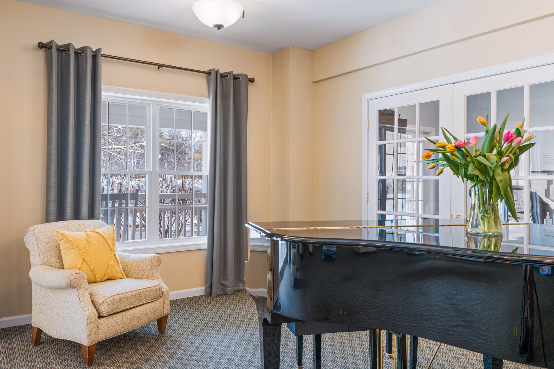 A cozy interior room with a patterned armchair featuring a yellow cushion, a large window with gray curtains, a black grand piano with a vase of colorful tulips on top, and white French doors in the background.