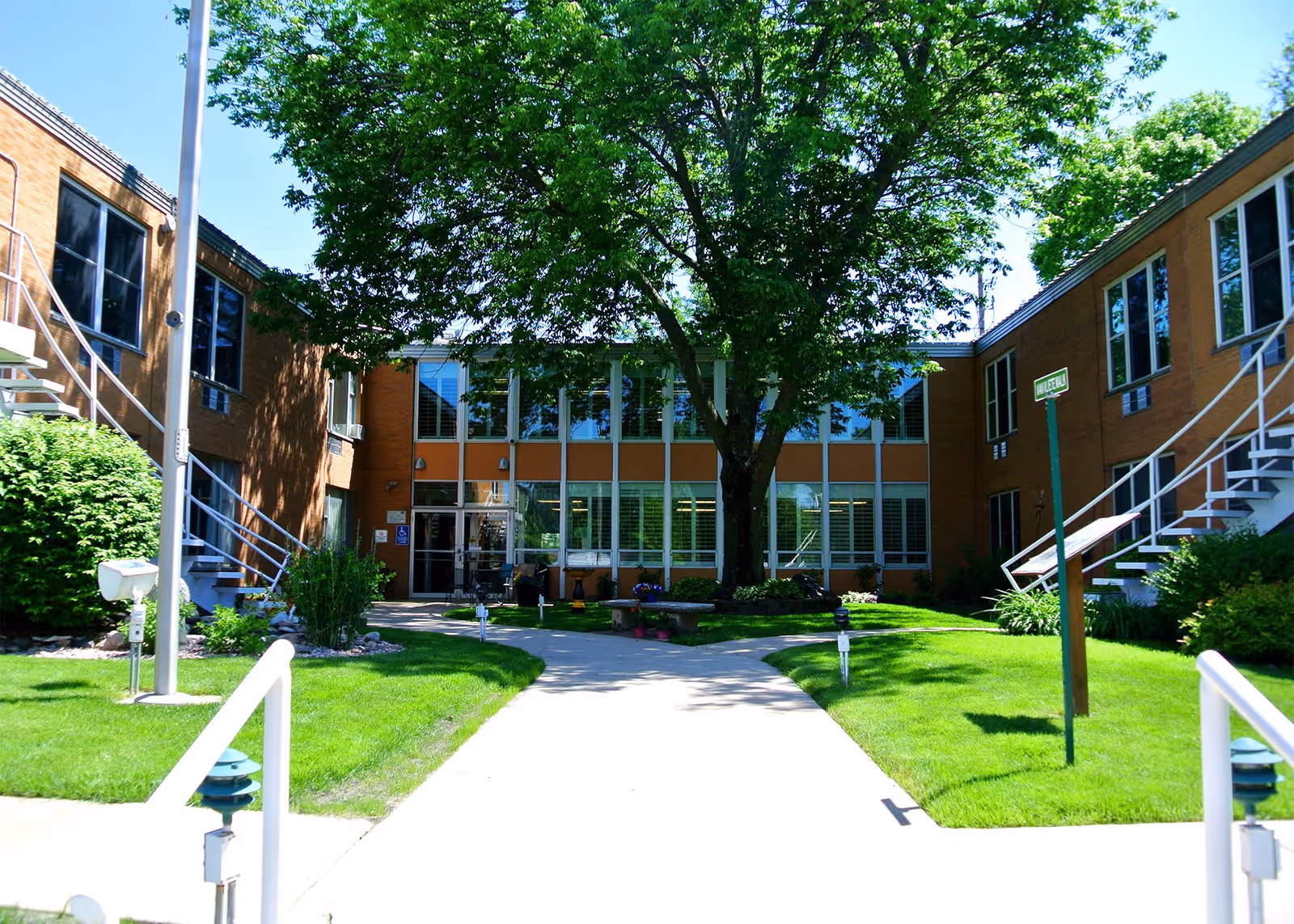 Outdoor courtyard area of King’s Gardens Campus of Perry Lutheran Homes featuring a large tree in the center, surrounded by green grass, bushes, and a paved walkway leading to a two-story brick building with large windows and exterior staircases on both sides.