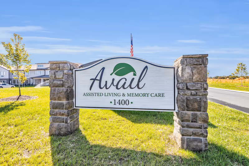 Entrance sign for Avail Assisted Living & Memory Care (1400) mounted between stone pillars on a grassy lawn with the facility and an American flag in the background.