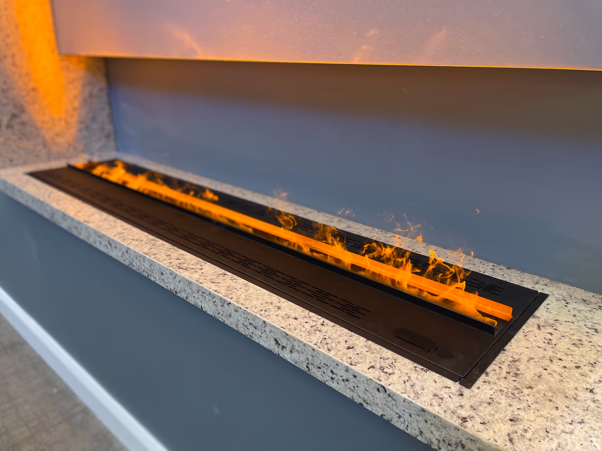 A modern, rectangular indoor fireplace with visible flames set into a speckled granite countertop against a gray wall.